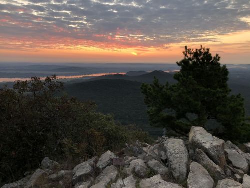 A scenic view of a sunset over rolling hills and a river, framed by rocky terrain and a lone tree.