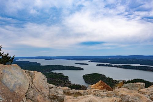 A scenic view of a lake surrounded by islands and greenery, with a cloudy sky above. Rocky foreground adds depth.