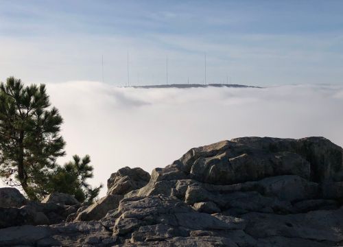A rocky landscape with a view of a fog-covered valley and radio towers emerging from the mist in the distance.