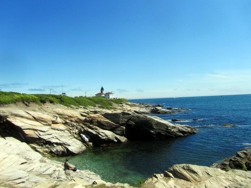 A rocky coastline with a clear blue sea and a lighthouse in the distance under a bright blue sky.