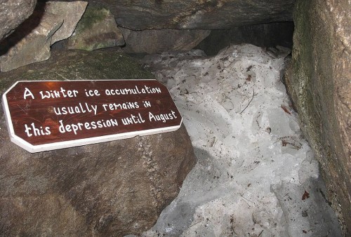 Sign in a rocky cave indicating a winter ice accumulation that lasts until August, with ice visible on the ground.