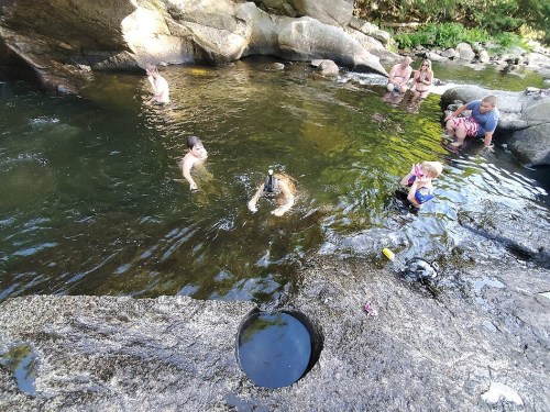A group of people enjoying a natural swimming area with rocks and clear water, some swimming and others relaxing nearby.
