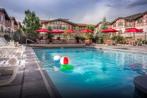 A sunny pool area with colorful beach balls, lounge chairs, and red umbrellas, surrounded by a fence and buildings.