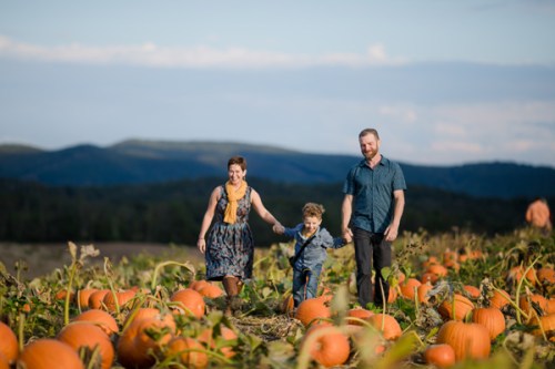 A family walks through a pumpkin patch, surrounded by large pumpkins and scenic hills in the background.