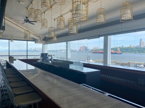 A modern bar with a marble countertop and hanging lights, overlooking a waterfront view with city skyline in the background.