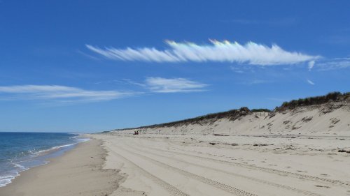 A serene beach scene with soft sand, gentle waves, and colorful clouds in a clear blue sky. Dunes line the shore.