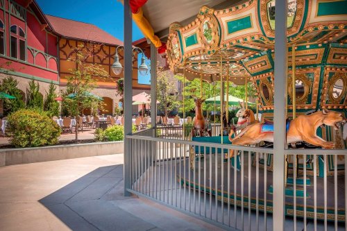 A colorful carousel with animal figures, surrounded by a patio with tables and umbrellas in a sunny outdoor setting.