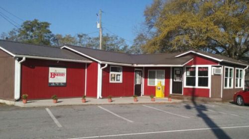 A red and brown building with a sign, surrounded by small plants, and parked cars in a sunny outdoor setting.