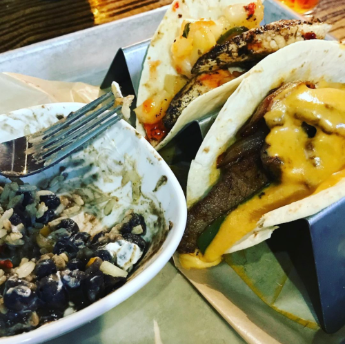Three tacos in a holder with melted cheese, alongside a bowl of black beans and rice, with a fork resting on the plate.