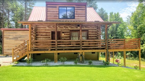 A rustic log cabin with a red metal roof, wooden deck, and green lawn surrounded by trees.