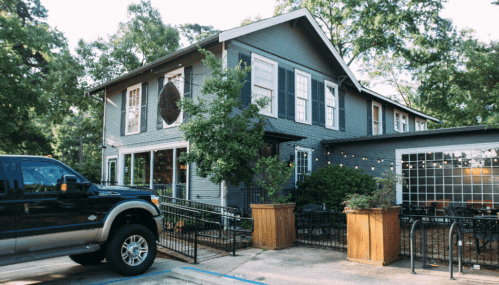A two-story gray house with large windows, surrounded by trees and outdoor seating, with a black truck parked nearby.