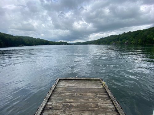 A wooden dock extends into a calm lake, surrounded by lush green trees under a cloudy sky.