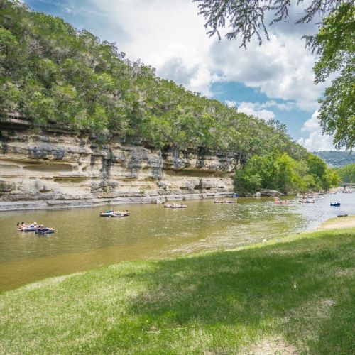 A serene river scene with people floating on tubes, surrounded by lush green cliffs and trees under a partly cloudy sky.
