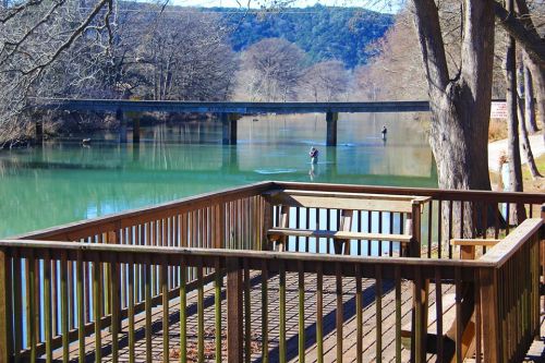A serene river scene with a wooden deck, trees, and a bridge in the background; two people fishing in the water.
