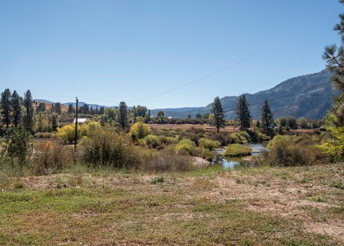 A serene landscape featuring a river, trees, and mountains under a clear blue sky.