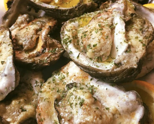 A close-up of baked oysters topped with herbs and a drizzle of sauce, served on a plate.