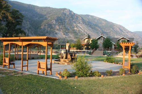 A scenic outdoor area with wooden pergolas, a fountain, and a mountain backdrop, featuring a spacious lawn and a house.