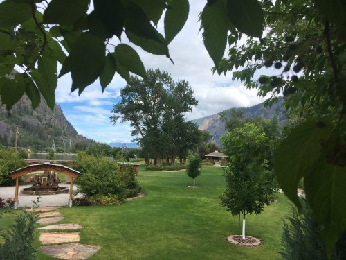 Lush green park with trees, pathways, and a scenic view of mountains and a lake under a cloudy sky.