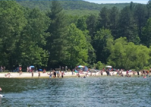 A busy beach scene with people swimming and relaxing under trees, surrounded by a calm lake and mountains in the background.