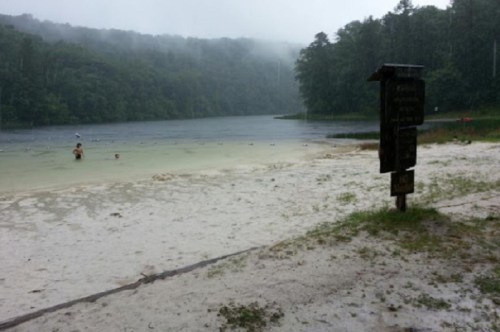 A rainy scene at a lake with a sandy beach, a signpost, and a few people in the water. Lush green trees in the background.