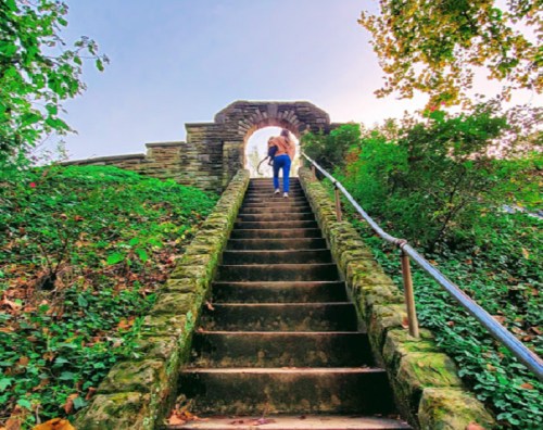 A person walks up stone steps surrounded by greenery, leading to an archway under a clear sky.