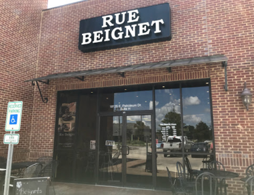 Exterior of Rue Beignet restaurant, featuring a brick facade, large windows, and an "OPEN" sign. Outdoor seating visible.