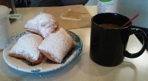A plate of powdered sugar-covered beignets next to a black coffee mug with a red straw.