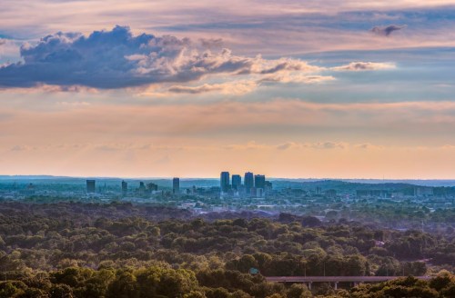 A panoramic view of a city skyline at sunset, surrounded by lush greenery and dramatic clouds in the sky.