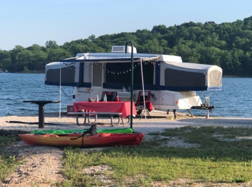 A camper trailer by a lake, with a picnic table and a colorful kayak in the foreground, surrounded by greenery.