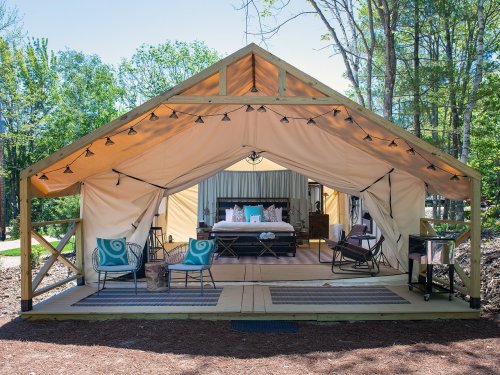 A cozy glamping tent with a bed, seating area, and string lights, set in a wooded outdoor environment.