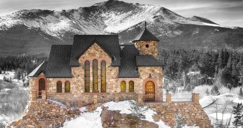 A stone church with large windows sits on a snowy hill, framed by a majestic mountain backdrop in black and white.