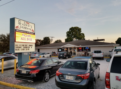 A seafood restaurant with a sign advertising all-you-can-eat shrimp and crab, surrounded by parked cars at sunset.