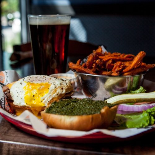 A plate with a burger topped with a fried egg, sweet potato fries, and a drink in a glass.