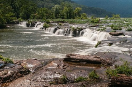 A serene river scene with cascading waterfalls over rocky terrain, surrounded by lush greenery and distant mountains.