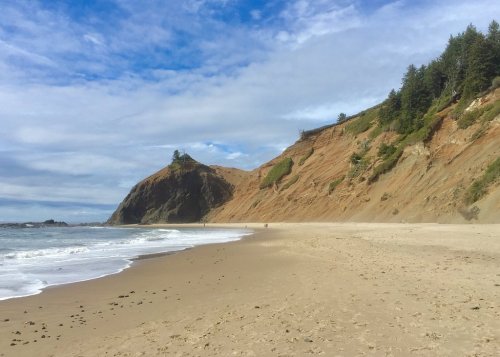 A sandy beach with gentle waves, flanked by steep cliffs and lush greenery under a partly cloudy sky.