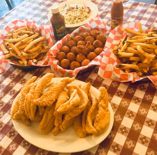 A table spread with fried fish, hushpuppies, French fries, coleslaw, and sauces in checkered baskets.