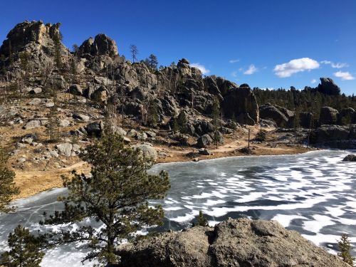A frozen lake surrounded by rocky terrain and pine trees under a clear blue sky.