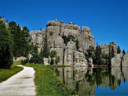 A scenic view of towering rock formations beside a calm lake, with a clear blue sky and lush greenery along the path.