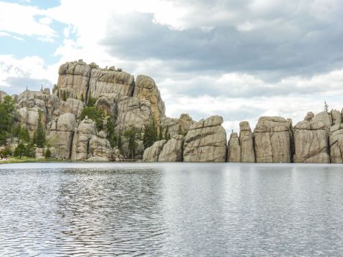 Rocky formations rise above a calm lake, surrounded by trees under a cloudy sky.