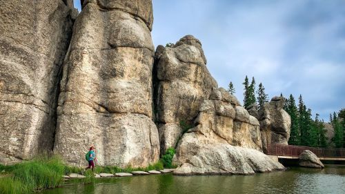 A hiker walks along a rocky shoreline beside a calm lake, surrounded by towering stone formations and pine trees.