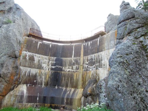 A tall dam surrounded by large rocks, with water cascading down its weathered surface. Green grass and wildflowers in the foreground.