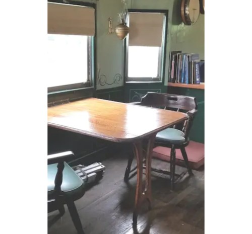 A wooden table with two chairs in a cozy room, featuring green walls and bookshelves. Natural light from windows.
