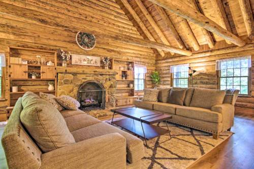 Cozy log cabin living room with a stone fireplace, wooden beams, and comfortable sofas. Natural light from large windows.