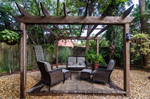 A wooden pergola with seating surrounded by greenery and gravel, creating a cozy outdoor relaxation area.