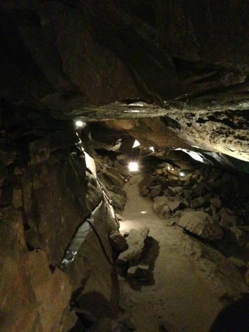 A dimly lit cave interior with rocky walls and a sandy path, illuminated by soft lights along the way.