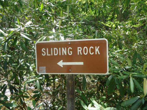 A brown sign with white text reading "SLIDING ROCK" and an arrow pointing left, surrounded by greenery.