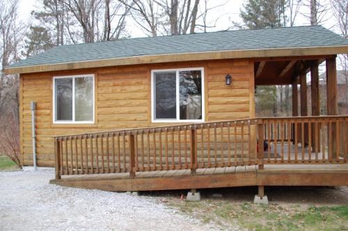 A wooden cabin with a ramp, large windows, and a porch, surrounded by trees and gravel pathway.