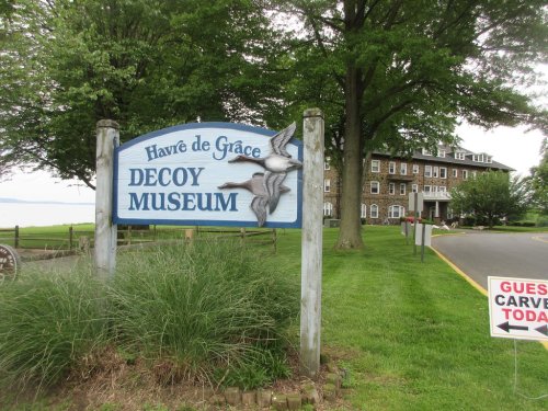 Sign for the Havre de Grace Decoy Museum, surrounded by greenery and a view of a historic building in the background.