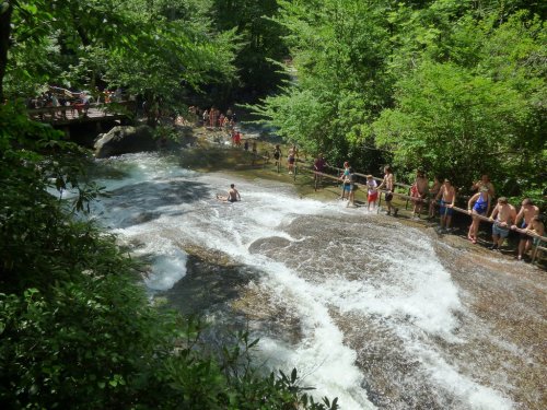 A busy natural swimming area with people enjoying the water and lush greenery surrounding a rocky stream.
