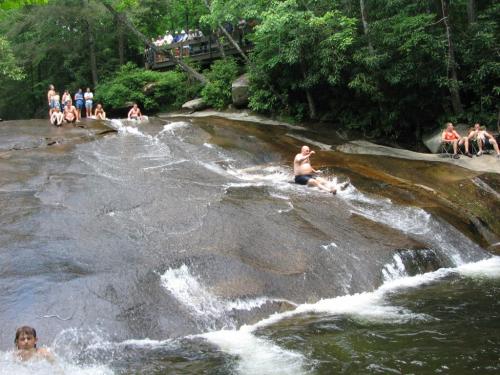 People enjoying a natural water slide, with some swimming and others watching from a rocky area surrounded by trees.